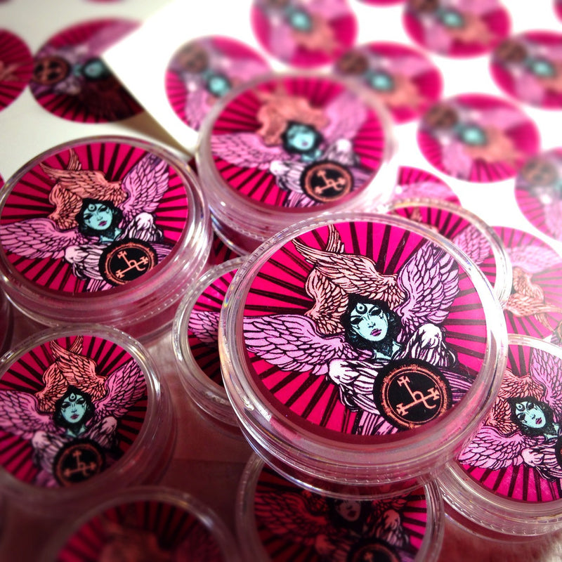 Round makeup jars with labels showing a black and pink drawing of a winged female goddess.