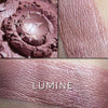 Lumine multi purpose illuminator loose and swatched on the skin. LUMINE: A deeper-toned, rosy plum with a soft blue iridescence.'