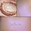OSTARA highlighter loose and swatched on the skin. Ostara is a soft peach with a strong purple highlight. Jar labels feature the art of Alphonse Mucha.