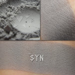 Syn matte eyeshadow shown loose and swatched on the skin. Soft grey with cool undertones.