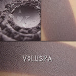 Voluspa matte eyeshadow shown loose and swatched on the skin. Medium-deep gray with strong violet/mauve tones.