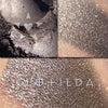 CLOTILDA eyeshadow shown loose and swatched on the skin. a rich, complex grey with strong gold highlights and multicolor iridescence.